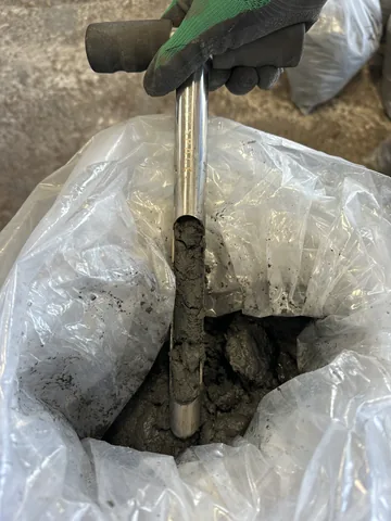 A gloved hand using a sampling tool inside a bag of material.